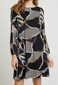 Svart klänning med kedja och mönstertryck med gyllene accenter. Långa ärmar och avslappnad passform. Tillverkad av mjukt, texturerat tyg.