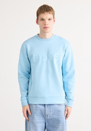 Young man wearing light blue sweatshirt with embossed "BOSS" logo and light blue jeans, standing against plain white background.