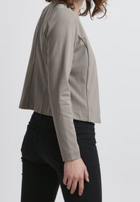 Kvinna iklädd en åtsittande taupe långärmad jacka och svarta jeans, sedd från sidan mot en enkel ljus bakgrund.