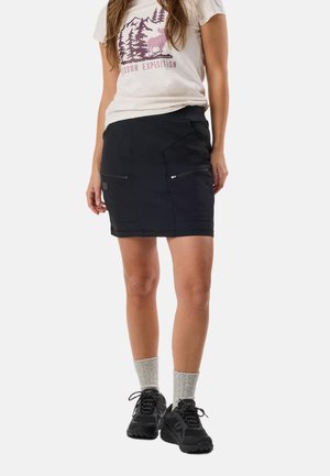 Woman wearing light beige graphic t-shirt, black zippered pocket skirt, gray socks, and black hiking shoes standing on white background.