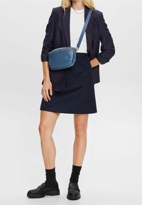 Jupe marine à coupe décontractée, associée à un blazer rayé. T-shirt blanc en dessous et sac bandoulière bleu. Mocassins noirs à semelle crantée.
