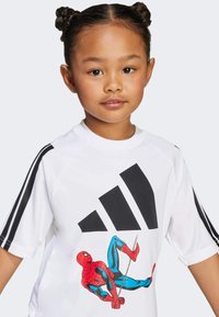 Biała koszula z krótkim rękawem z dużym czarnym logo Adidas i kolorową grafiką Spider-Mana. Zawiera czarne paski na rękawach.