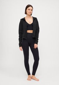 Fekete sportos összeállítás, amely egy cipzáras kapucnis felsőt, sportmelltartót és magasderékú leggingset tartalmaz sima anyagból. Egyszerű dizájn, minimális kiegészítők.