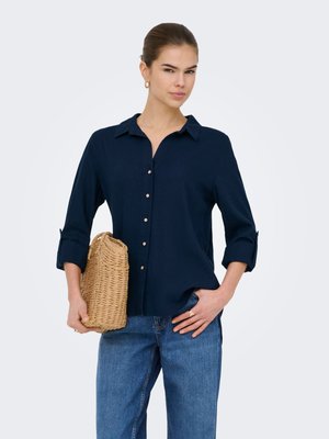 Femme portant une chemise bleu marine boutonnée et un jean bleu tenant une pochette tissée beige, cheveux tirés en arrière et boucles d’oreilles créoles dorées.