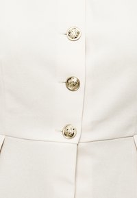 Τρία χρυσά διακοσμητικά κουμπιά τοποθετημένα κάθετα σε λευκό υφασμα με υφή, πιθανώς μέρος ενός ενδύματος.