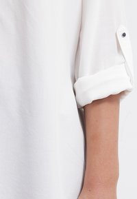 Blouse blanche avec des manches retroussées, fabriquée en tissu léger. Présente un bouton sur la manche et une texture lisse.