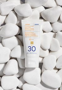 Crema viso solare colorata in un tubetto bianco; presenta SPF 30, protezione UVA/UVB e informazioni sul prodotto in testo arancione e blu, posizionata su pietre bianche.