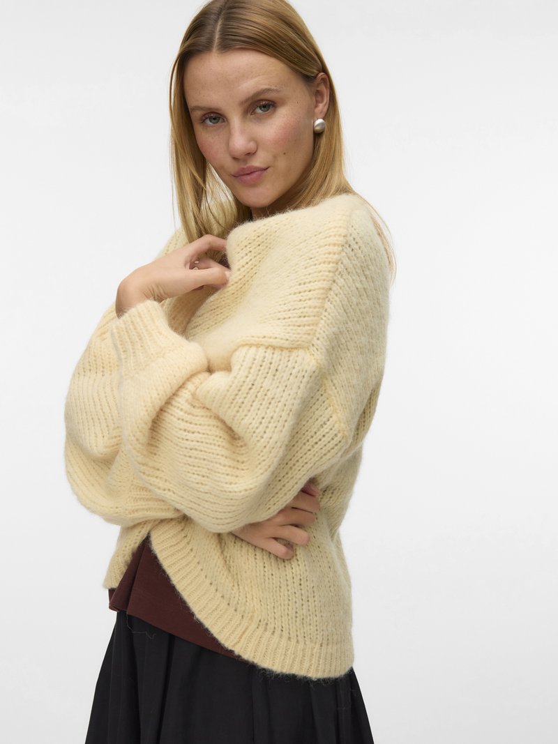 Blonde Frau mit großen Perlenohrringen, einem übergroßen cremefarbenen Strickpullover über einem braunen Oberteil und einem schwarzen Rock, die vor weißem Hintergrund posiert.