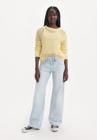 Pull en maille jaune clair avec un motif texturé, assorti à un jean bleu clair à jambes larges et des baskets noires en toile.