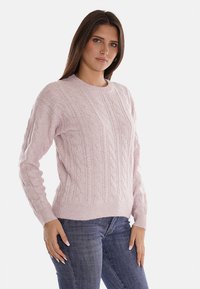 Maglione lavorato a maglia di un rosa chiaro con motivo a trecce, scollatura rotonda e polsini e orlo a coste. Abbinato a jeans blu scuro.