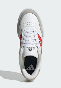 Bijele kožne tenisice s crvenim i crnim detaljima, zamotani vrh od antilopa, perforirani detalji na gornjem dijelu i teksturirana jezičak. Prisutna je Adidasova oznaka.