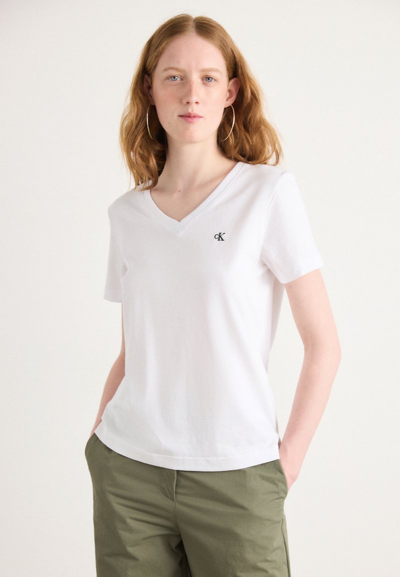 Maglietta di cotone bianca con scollo a V, maniche corte e un piccolo logo nero sul lato sinistro del petto. Indossata con pantaloni verdi, larghi.