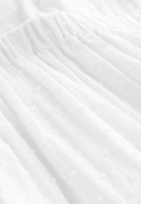 Tissu blanc avec un design texturé présentant des points en relief. Le matériau est léger et transparent, froncé en haut avec un élastique.