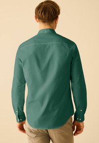 Cămașă lungă cu mânecă închisă la culoare, de un verde închis, cu guler cu nasturi, textură netedă, având accente cu nasturi la manșete și o margine curbată.