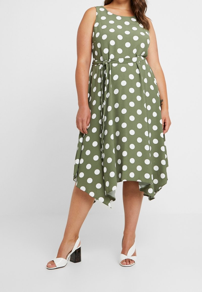 Grünes ärmelloses Kleid mit weißen Polka-Punkten, A-Linie-Silhouette, tailliert und asymmetrischem Saum; kombiniert mit weißen Blockabsatzsandalen.