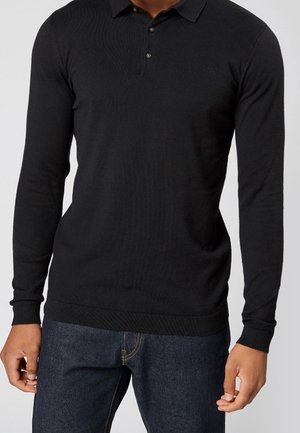 Man wearing a fitted black long-sleeve polo shirt paired with dark blue jeans, standing with arms relaxed by his sides.