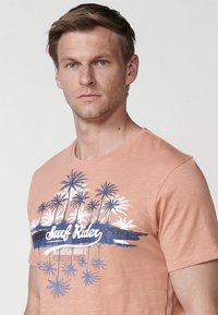 Īsām piedurknēm korallkrāsas T-krekls, izgatavots no mīksta auduma, ar baltu un zilu palmu koku grafiku centrā ar uzrakstu "Surf Rider".
