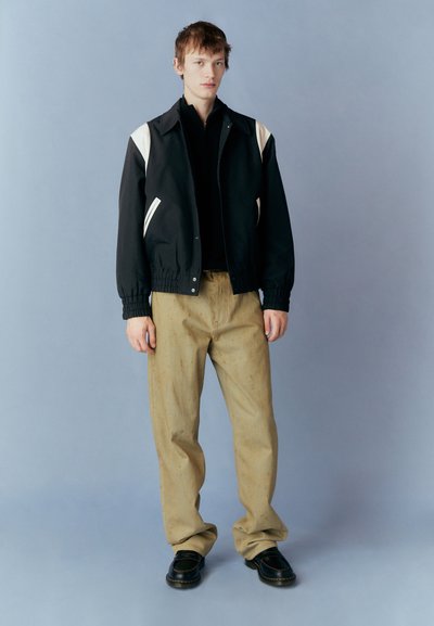 Black bomber jacket with cream accents, relaxed fit, elastic cuffs, paired with beige textured trousers and black loafers on a light background.