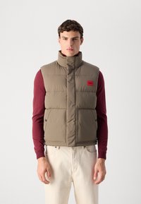 Young man wearing beige puffer vest over maroon long-sleeve shirt and off-white pants, standing against plain light background.