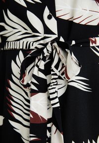 Black fabric with a white and burgundy leaf pattern. Features a tied waist belt and a button closure, emphasizing its structured design.