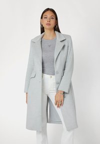 Cappotto in lana grigio chiaro con colletto a punta, bottoni monopetto, tasche laterali e taglio sartoriale, abbinato a un top grigio e pantaloni bianchi.