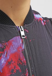 Svart bomberjacka med ett färgglatt abstrakt blommönster i röd och lila. Silverdragkedja med en märkesdragrest och ribbad krage.