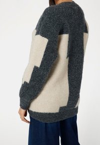 Pull tricoté en beige et gris foncé, avec des motifs géométriques en blocs, un col rond, des poignets et une base côtelés. Texture douce.