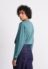 Chemise longue à manches longues turquoise avec un col rond, dotée d'une coupe décontractée. Associée à un jean bleu foncé, mettant en avant un design simple.