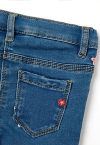Jeans de denim azul con un pequeño bolsillo trasero adornado con bordados florales en rosa y azul. La costura es visible a lo largo de las costuras.