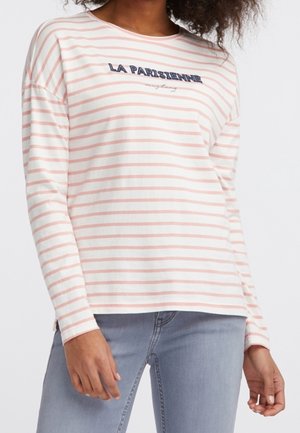 Žena nosící bílou košili s dlouhým rukávem, růžovými pruhy a nápisem "LA PARISIENNE", ve spojení s světle modrými džínami.