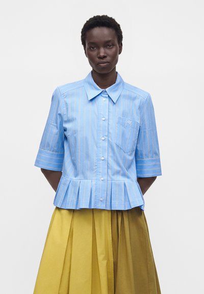 Femme portant une chemise bleu clair à manches courtes boutonnée avec des rayures blanches et une jupe plissée jaune, debout devant un fond uni.