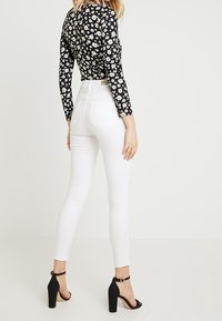 Top negro de manga larga con patrón floral blanco, combinado con jeans blancos ajustados y sandalias negras de tacón alto, visto desde atrás.