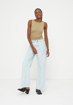 Femme debout devant un fond blanc, portant un haut beige sans manches, un jean large bleu clair et des chaussures à talons noires.