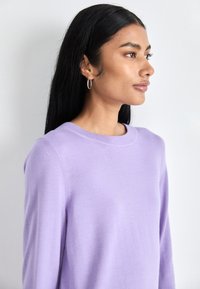 Sweater de malha lavanda com decote redondo, mangas ajustadas e uma textura suave. Modelo usa brincos de argola prateados.