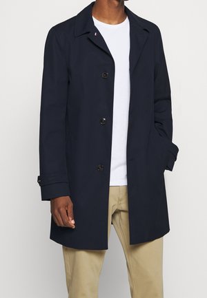 Mann trägt marineblauen, geknöpften Mantel über weißem T-Shirt und beigen Hosen, steht mit einer Hand in der Manteltasche vor einfarbigem Hintergrund.