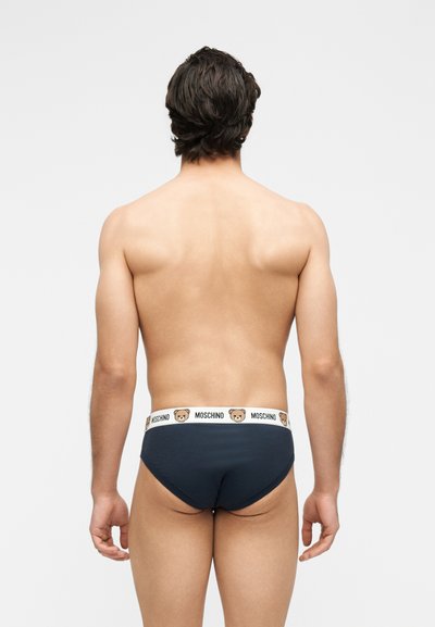 Άνδρας με σκούρα μαλλιά που φοράει μαύρα εσώρουχα με λευκή ζώνη που φέρει πρόσωπα αρκουδάκια και το κείμενο "MOSCHINO", εμφανιζόμενος από πίσω.