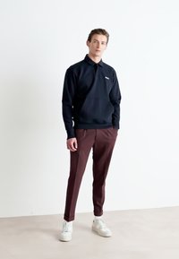 Polo preto de mangas longas, com gola canelada, combinado com calças numa tonalidade bordeaux escuras e tênis brancos. Design limpo e corte moderno.