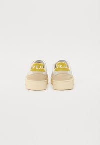 Sneaker bianche e beige con finitura in suede, dotate di un logo giallo sul tallone e una suola testurizzata per una migliore aderenza.