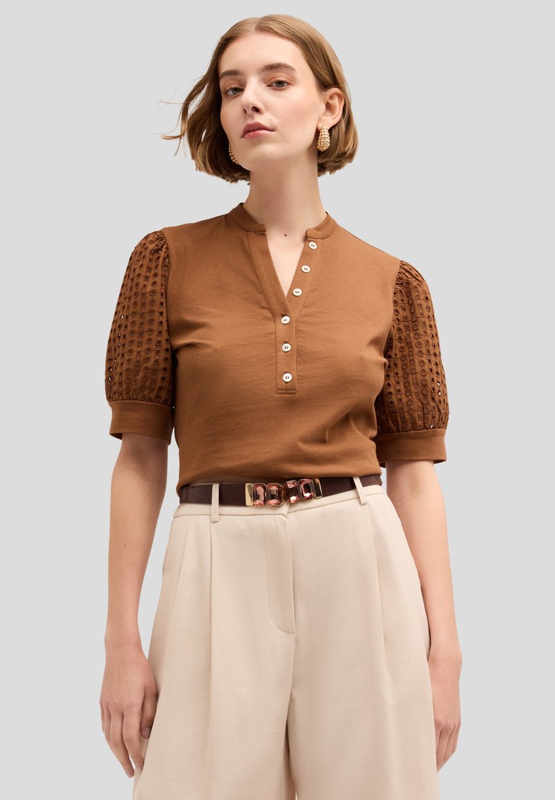 Braunes Oberteil mit Henley-Ausschnitt und kurzen Puffärmeln, das ein strukturiertes Muster aufweist. Kombiniert mit cremefarbenen, hochgerüschten Hosen und einem Gürtel.