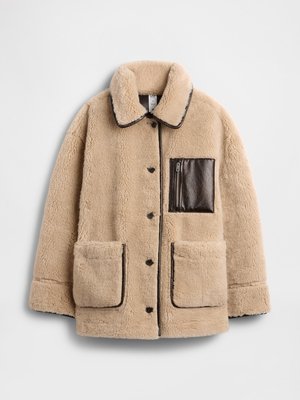 Casaco sherpa bege com um bolso com fecho em pele castanha, grandes bolsos frontais e gola, apresentando acabamentos e botões escuros em contraste.
