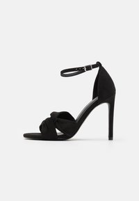 Svart mocka högklackad sandal med en knuten framsida, öppen tå, ankelrem med spänne och en slank stilettklack.