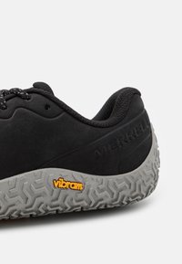 Schwarzer Schuh mit Obermaterial aus Wildleder und Stoff, strukturierten Gummisohle mit geometrischem Muster und Vibram-Logo in Orange an der Seite.