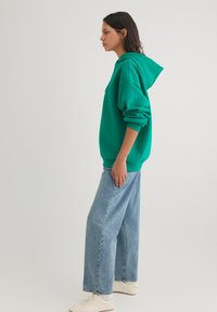 Green hoodie with a spacious fit and a front pocket, paired with light blue wide-leg jeans and beige sneakers. Simple, minimalistic design.