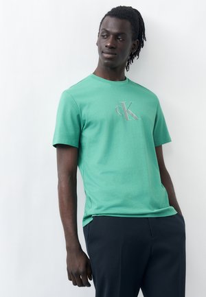 Άνδρας με σκουρόχρωμο δέρμα και dreadlocks, που φορά πράσινο t-shirt με το λογότυπο CK και σκούρο παντελόνι, στέκεται μπροστά σε απλό λευκό φόντο.