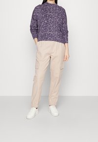 Femme debout portant un pull violet à imprimé animal, un pantalon cargo beige avec poches et des baskets blanches sur un fond clair uni.
