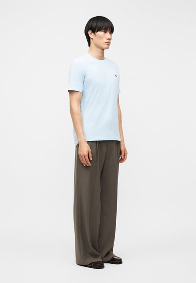 Άνδρας που στέκεται φορώντας ανοιχτό μπλε πουκάμισο με κοντά μανίκια και μικρό λογότυπο, φαρδιά καφέ παντελόνια και σκούρα καφέ παπούτσια σε απλό λευκό φόντο.