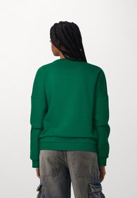 Sudadera verde con escote redondo y hombros caídos, hecha de una tela suave; combinada con pantalones cargo grises que cuentan con bolsillos laterales.