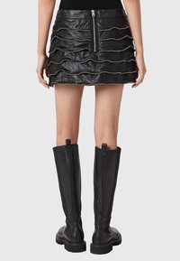 Γυναίκα που φοράει μαύρη δερμάτινη μίνι φούστα με οριζόντιες στρώσεις φερμουάρ και μαύρες μπότες ως το γόνατο, απεικονίζεται από πίσω.