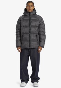 Grå puffjacka med huva, med dragkedja fram, två sidofickor och kviltad design. Matchas med mörka denimbyxor och vita skor.