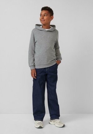 Chłopiec stoi na jednolitym tle, ubrany w szary hoodie, ciemnoniebieskie spodnie i białe trampki, z jedną ręką w kieszeni.
